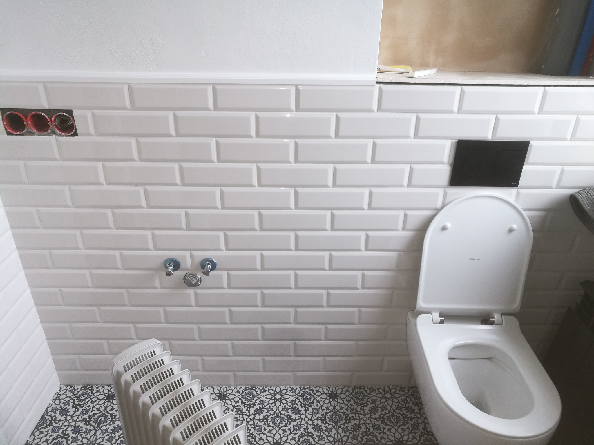 Biała toaleta kompaktowa z otwartą klapą na tle ściany z białych, błyszczących płytek typu metro, widoczne instalacje wodne i elektryczne, podłoga wyłożona płytkami z wzorem.