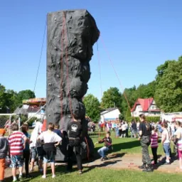 Mobilna ścianka wspinaczkowa na trawiastym terenie, z grupą dzieci i młodzieży obserwujących wspinaczkę, w tle budynki i wóz strażacki.