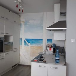 Wawerscy Malarze - Biała kuchnia z nowoczesną zabudową, na ścianie fototapeta z motywem plaży i morza, widoczne narzędzia i butelka wody na blacie.