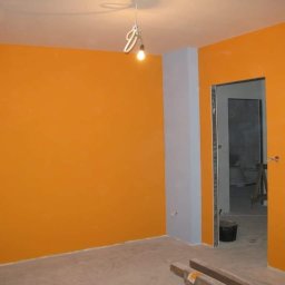 Wawerscy Malarze - Pomarańczowe ściany świeżo po malowaniu, szara kolumna przy wejściu, surowy sufit z wiszącą żarówką, wiadro i drewniane listwy na podłodze.