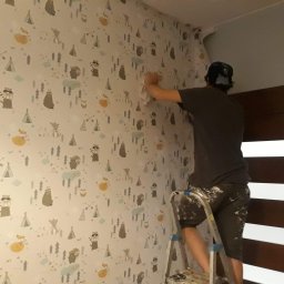 Wawerscy Malarze - Mężczyzna na drabinie wygładza białą tapetę w dziecięcym pokoju, pokrytą wzorami zwierząt, tipi i drzew na neutralnym tle, widoczny fragment odklejającej się tapety przy suficie.