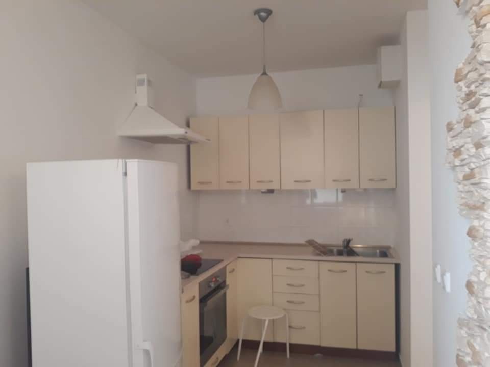 Jasna kuchnia z beżowymi szafkami, białym sprzętem AGD i dekoracyjną ścianą z kamienia, widok na mały stołek barowy.