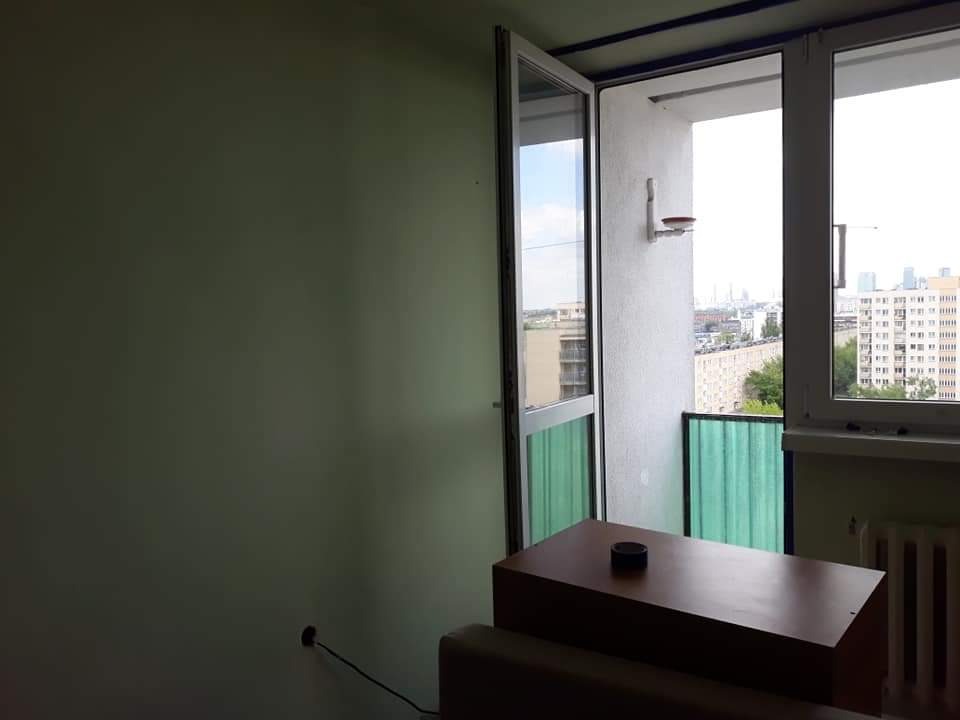 Pokój z zieloną ścianą przed balkonem z widokiem na miasto, widoczne gniazdko elektryczne i grzejnik.