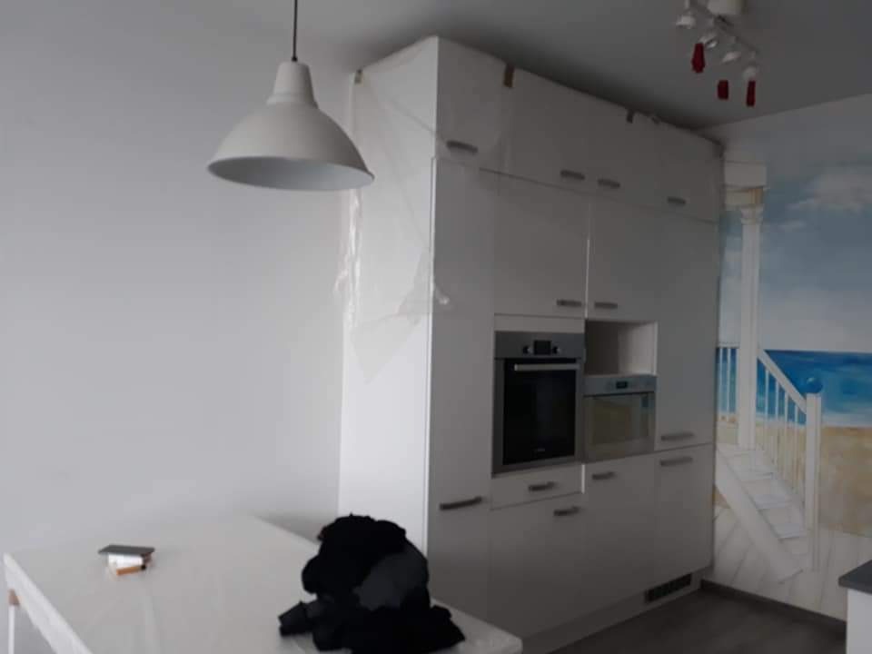 Biała kuchnia w trakcie zabezpieczania folią przed pracami wykończeniowymi, widoczna lampa wisząca i fragment muralu z motywem morskim.
