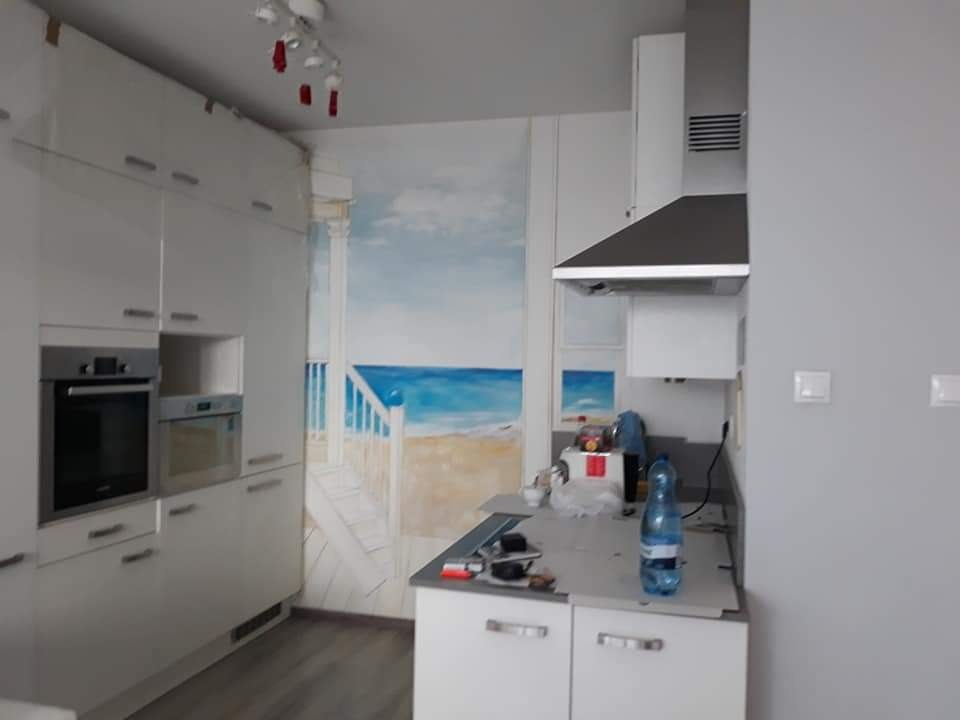 Biała kuchnia z nowoczesną zabudową, na ścianie fototapeta z motywem plaży i morza, widoczne narzędzia i butelka wody na blacie.