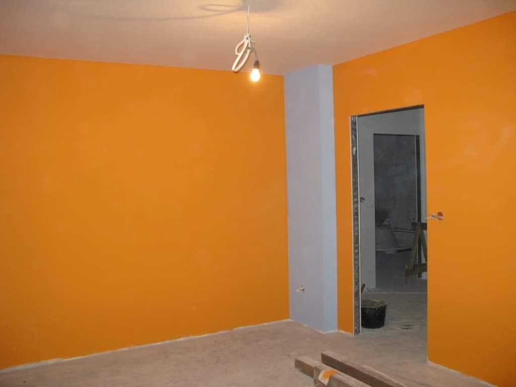 Pomarańczowe ściany świeżo po malowaniu, szara kolumna przy wejściu, surowy sufit z wiszącą żarówką, wiadro i drewniane listwy na podłodze.