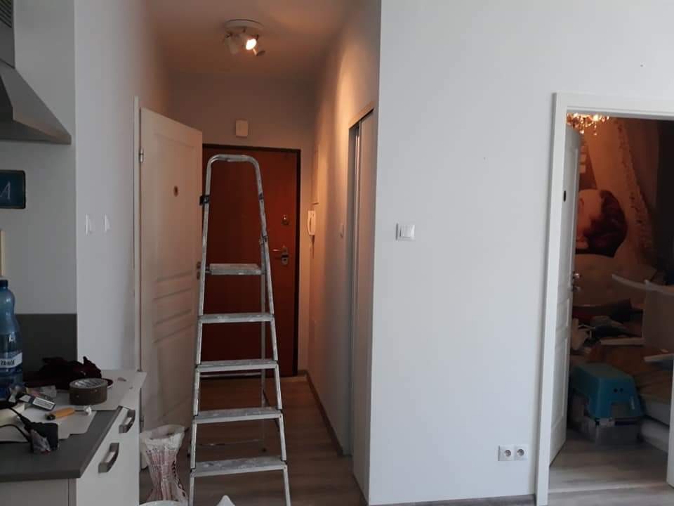 Korytarz w trakcie odświeżania: świeżo pomalowane na biało ściany, aluminiowa drabina rozstawiona w przejściu, widoczne ślady prac malarskich na podłodze i elementach wyposażenia.