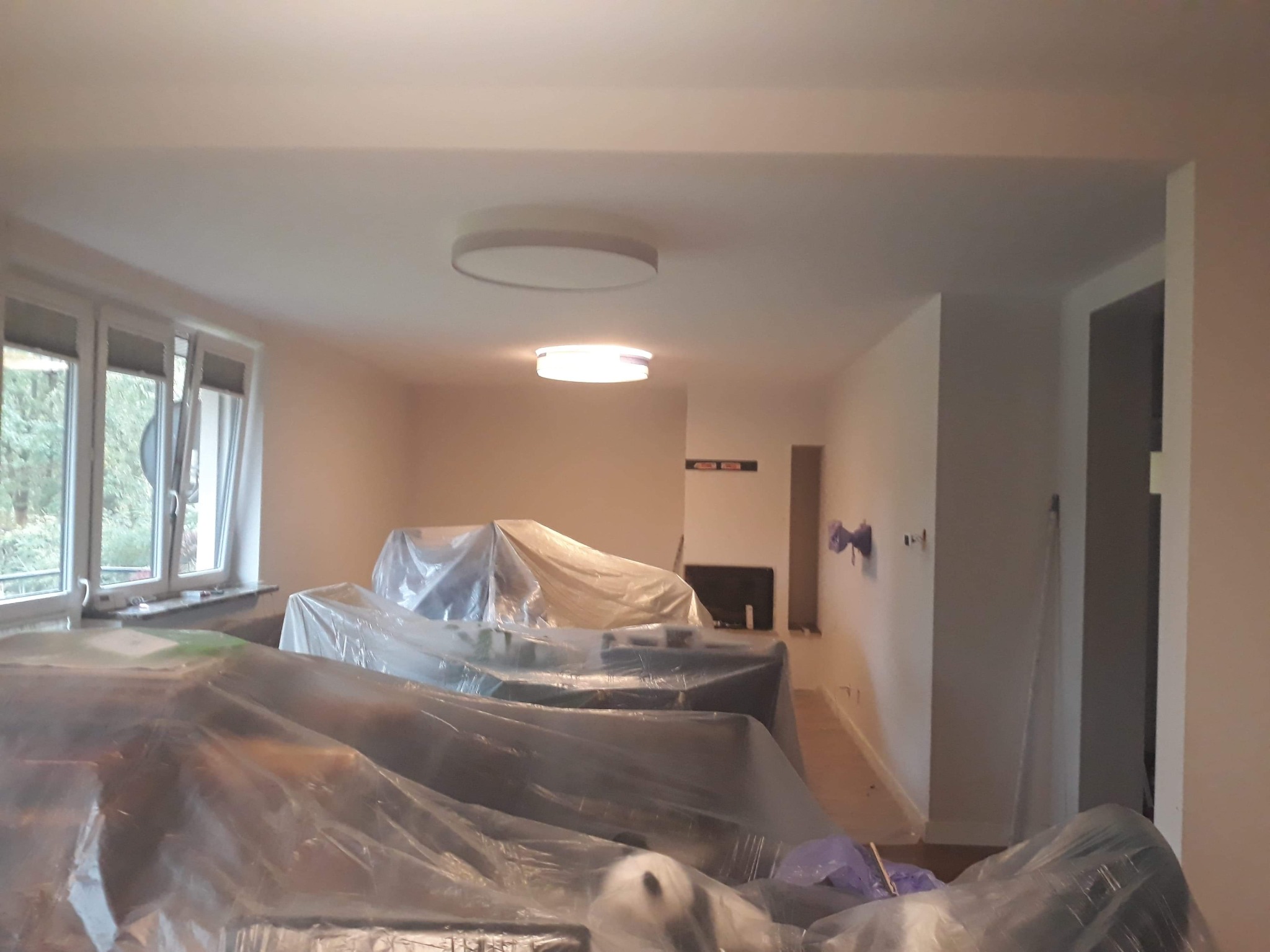 Pokój z meblami zabezpieczonymi folią malarską przed rozpoczęciem prac wykończeniowych, widoczne okna i oświetlenie sufitowe.