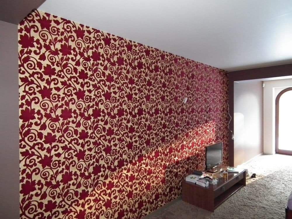 Ściana w pokoju pokryta tapetą w motywy roślinne w kolorze czerwonym i beżowym, widoczny fragment pokoju z telewizorem i drzwiami.