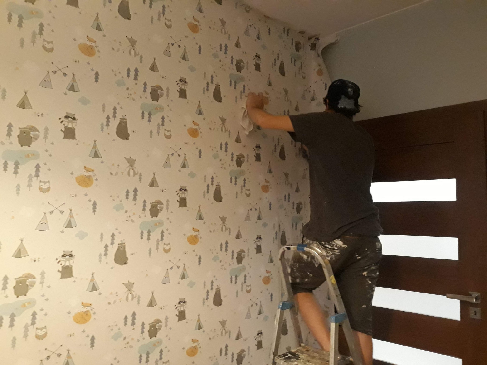Mężczyzna na drabinie wygładza białą tapetę w dziecięcym pokoju, pokrytą wzorami zwierząt, tipi i drzew na neutralnym tle, widoczny fragment odklejającej się tapety przy suficie.