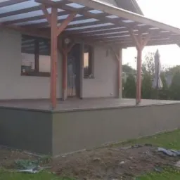 Zadaszony taras z drewnianą konstrukcją wspartą na słupach, widoczny z poziomu trawnika, z elementami wykończenia w kolorze szarości.