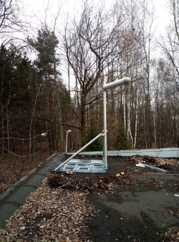 Instalacja odgromowa z uchwytem na dachu budynku, widoczne mocowania, tło stanowią drzewa bez liści.