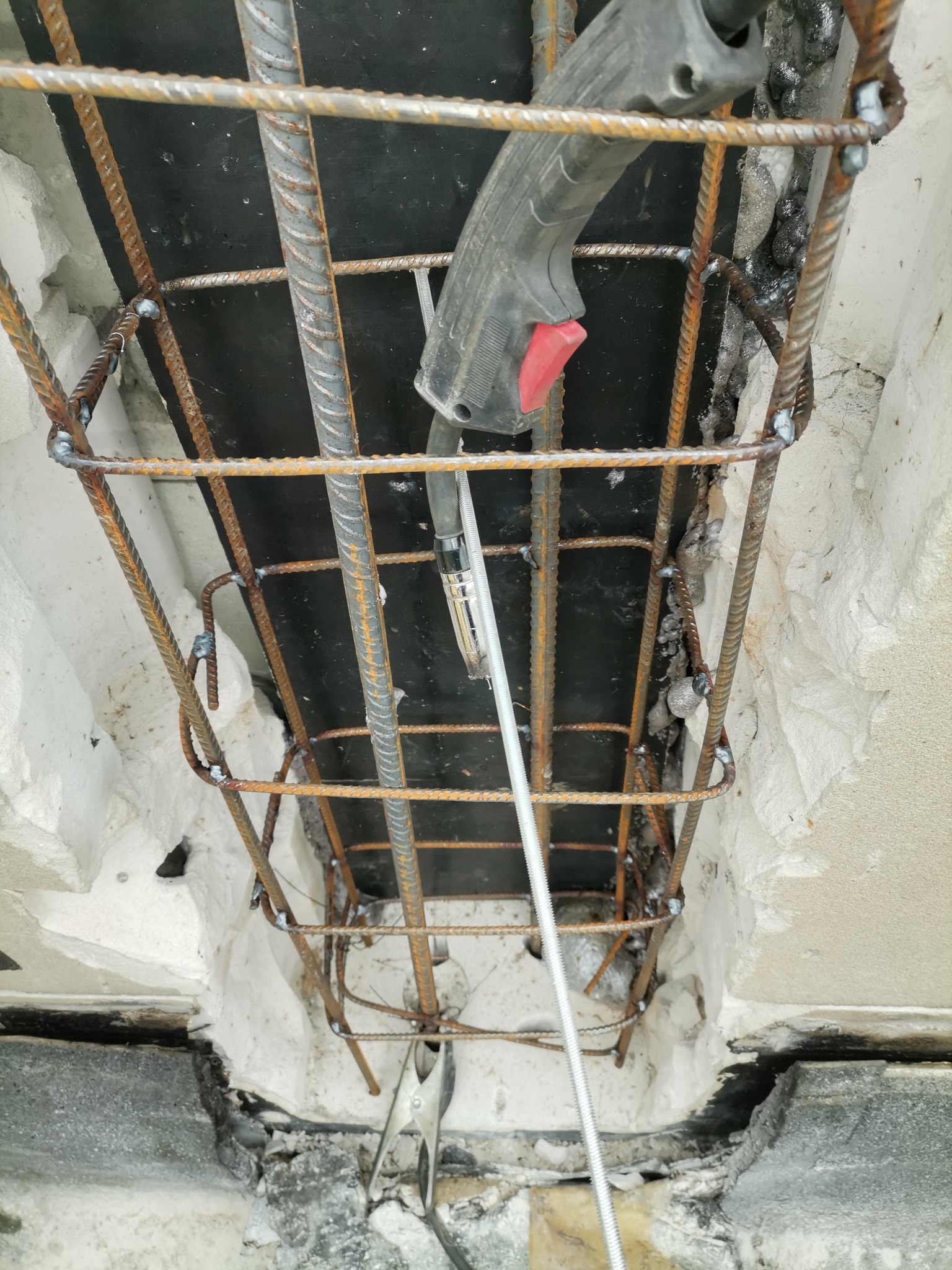 Zespawane zbrojenie z prętów żebrowanych, widoczny uchwyt spawarki z czerwonym przyciskiem i przewód, całość osadzona w betonowej konstrukcji z ubytkami.