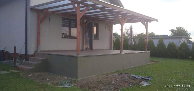 Zadaszony taras z drewnianą konstrukcją wspartą na słupach, widoczny z poziomu trawnika, z elementami wykończenia w kolorze szarości.