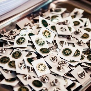 Kolekcja przypinek z logo adwokackim na białych kartonikach, ułożonych w metalowej tacy. Widoczne detale logo: zielony wzór na złotym tle. Różne ułożenia przypinek tworzą wzór.