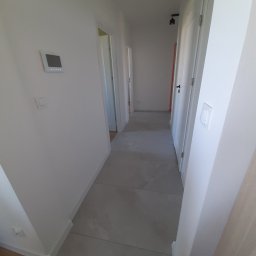 Usługi ogólnobudowlane Budo-styl Piotr Korenkiewicz - Jasny korytarz z szarą podłogą z płytek i białymi ścianami z widocznym panelem sterowania. W tle widoczne drzwi.