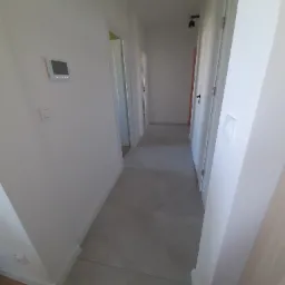 Jasny korytarz z szarą podłogą z płytek i białymi ścianami z widocznym panelem sterowania. W tle widoczne drzwi.