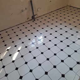 Usługi ogólnobudowlane Budo-styl Piotr Korenkiewicz - Biało-czarna podłoga z płytek ułożonych w karo, świeżo po fugowaniu. Widoczne ślady po remoncie na ścianach i rura przy ścianie. Całość w jasnym oświetleniu.