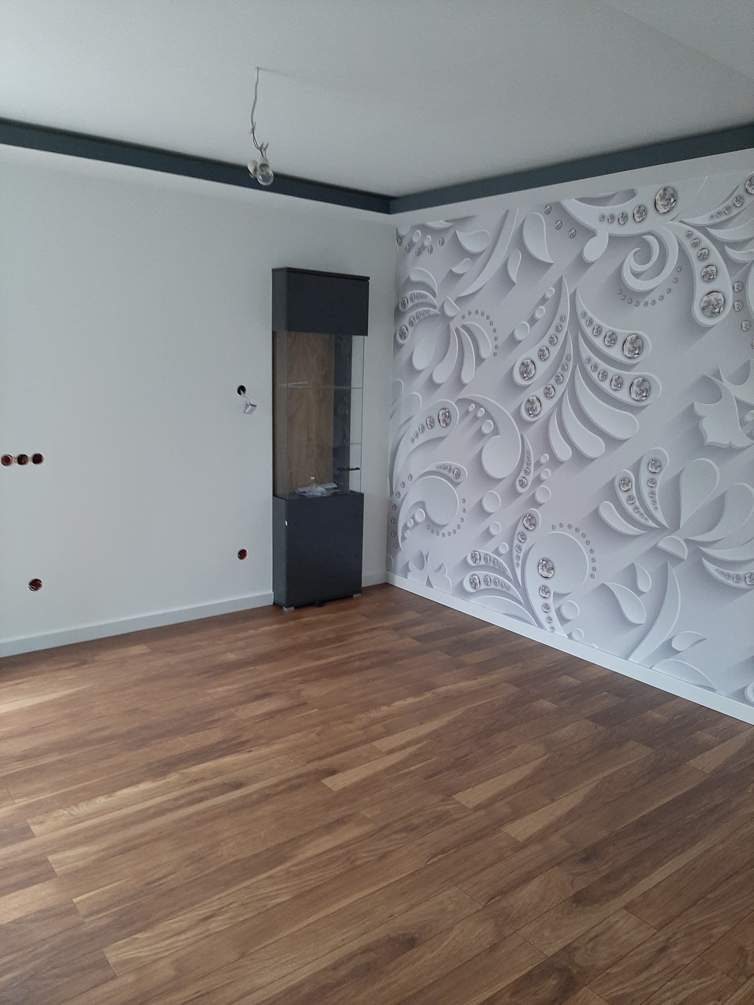 Pokój po remoncie z panelami podłogowymi w odcieniach brązu, ściana z dekoracyjną tapetą w odcieniach bieli i szarości z motywem roślinnym i kryształkami, oraz szara listwa pod sufitem i czarna...