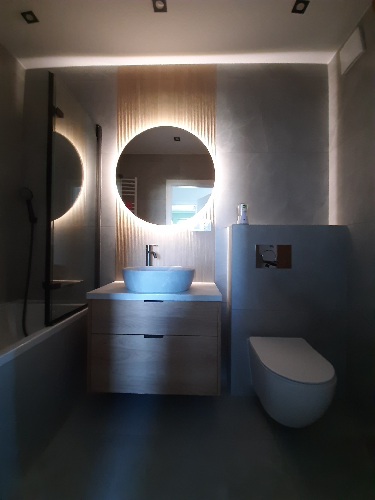 Nowoczesna łazienka w szarościach z okrągłym lustrem podświetlanym LED i drewnianą szafką umywalkową. Widoczna toaleta podwieszana i fragment kabiny prysznicowej.