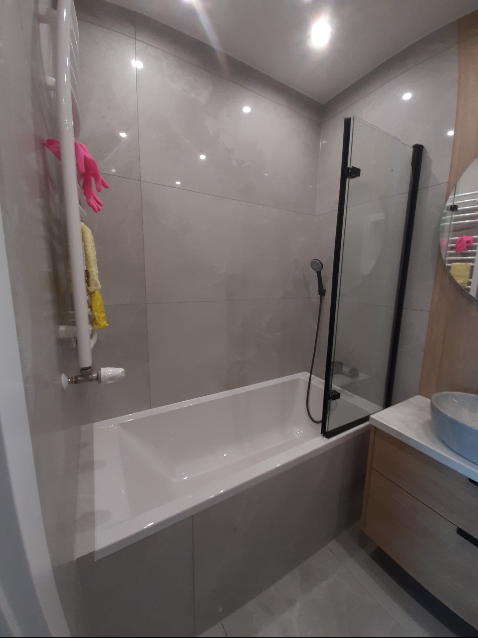 Nowoczesna łazienka z wanną i szklanym parawanem, szare płytki imitujące marmur, grzejnik drabinkowy z rękawicami i ściereczką, umywalka nablatowa.