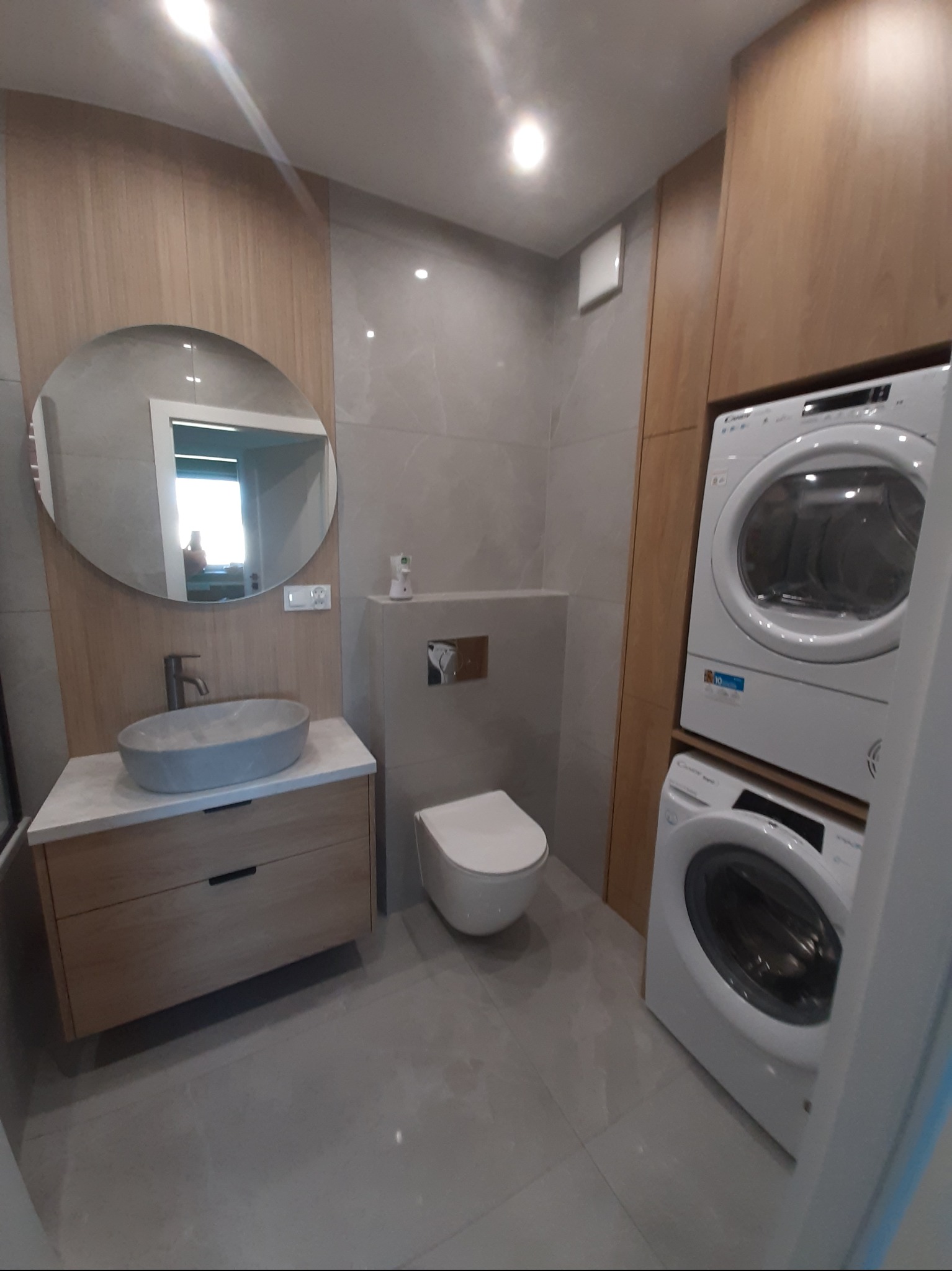 Jasna łazienka z okrągłym lustrem, kamienną umywalką i zabudowaną pralko-suszarką. Minimalistyczny design, szare płytki i drewniane akcenty tworzą przytulną przestrzeń.