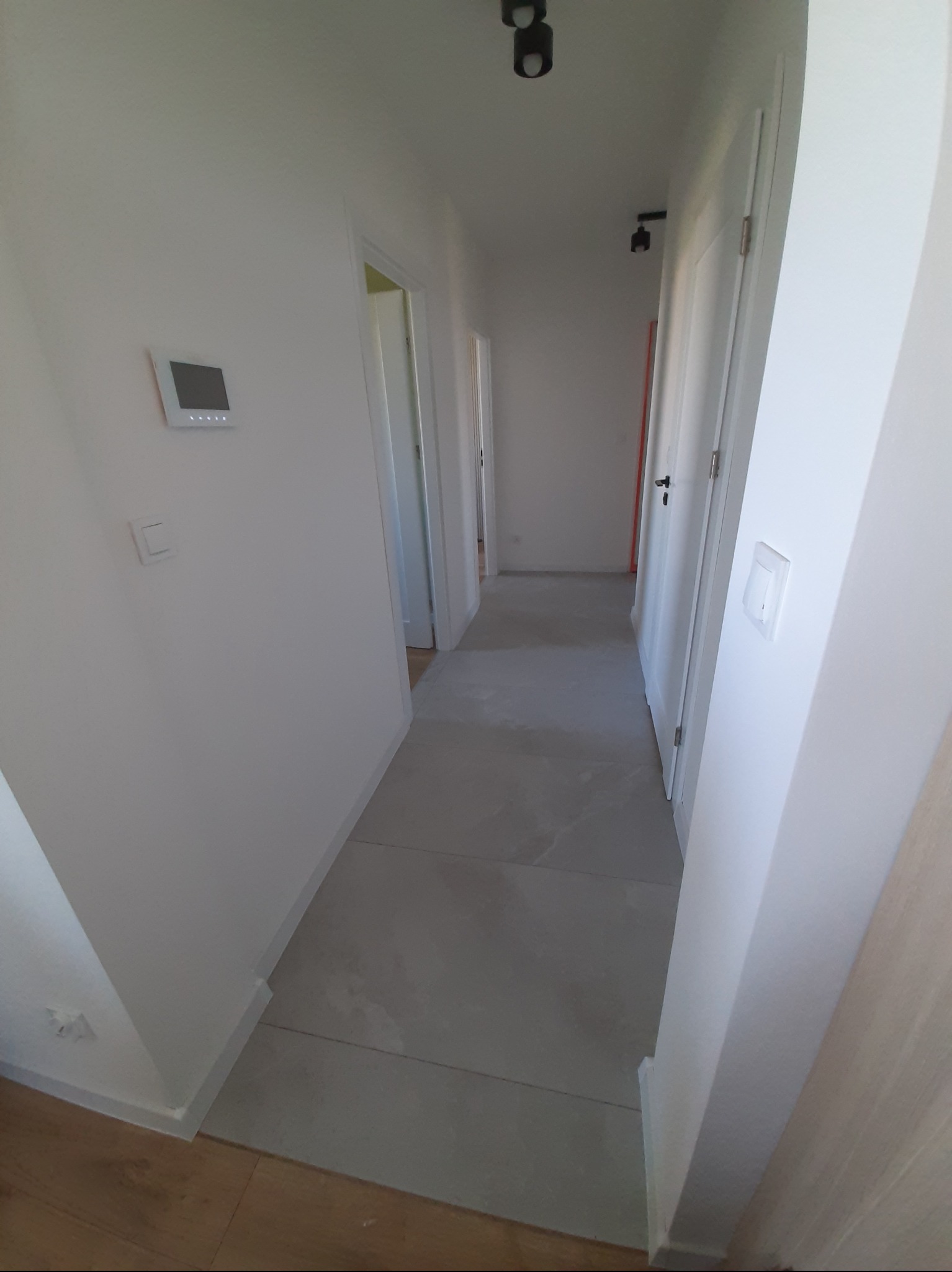 Jasny korytarz z szarą podłogą z płytek i białymi ścianami z widocznym panelem sterowania. W tle widoczne drzwi.