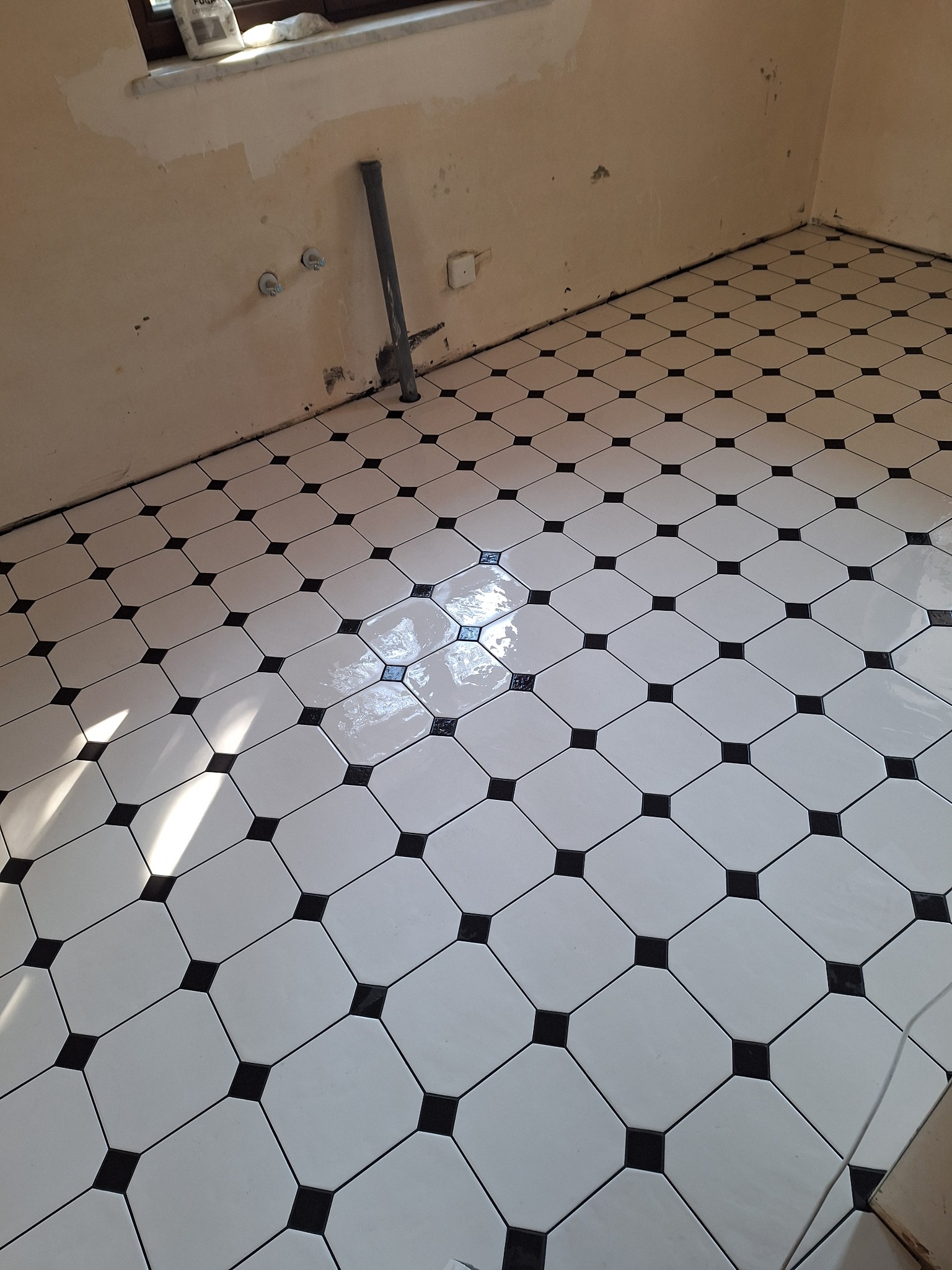 Biało-czarna podłoga z płytek ułożonych w karo, świeżo po fugowaniu. Widoczne ślady po remoncie na ścianach i rura przy ścianie. Całość w jasnym oświetleniu.