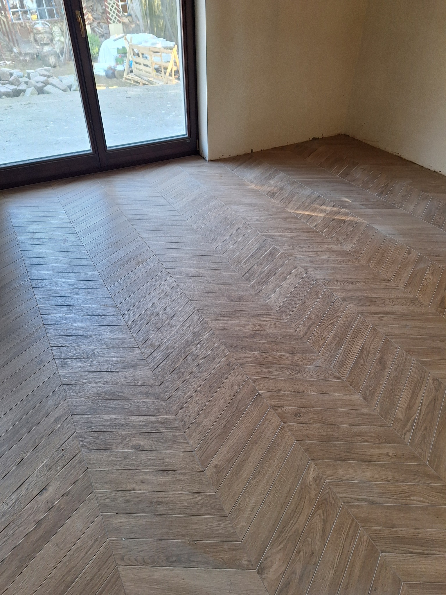 Ukończona podłoga z płytek imitujących drewno, ułożonych we wzór jodełki francuskiej, widoczna przy oknie w surowym wnętrzu. Jasne, naturalne światło.