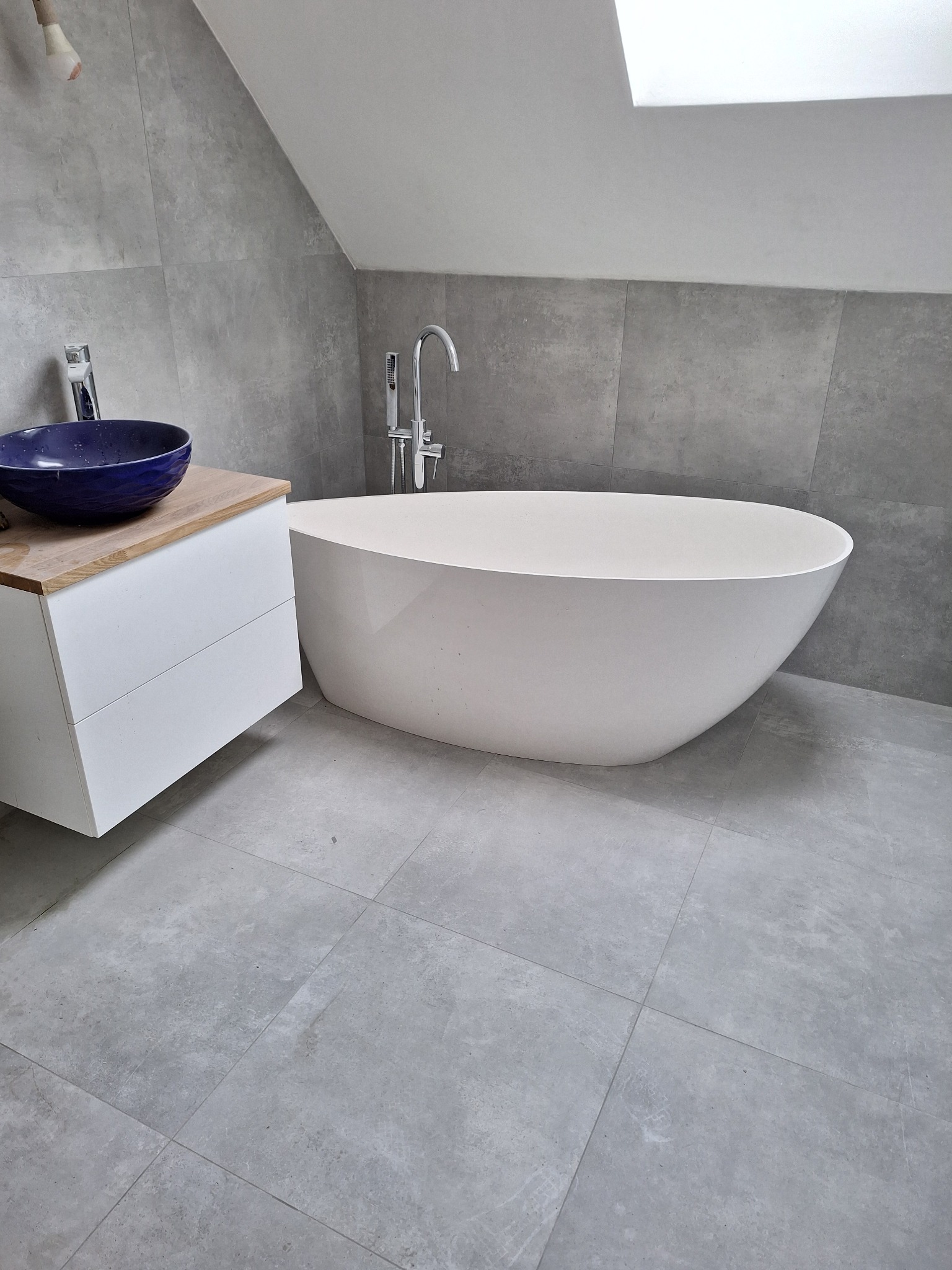 Nowoczesna łazienka w szarościach: wolnostojąca wanna, umywalka nablatowa w odcieniach granatu i drewniany blat. Minimalistyczny design, płytki imitujące beton.
