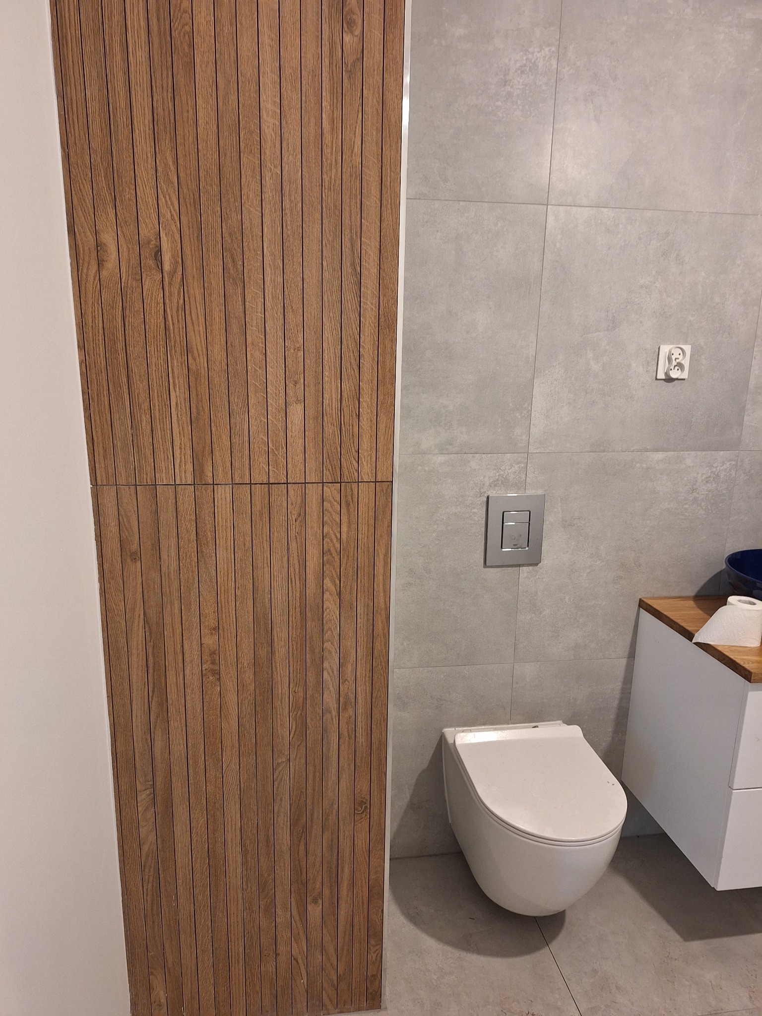 Nowoczesna łazienka z drewnianą zabudową, szarą płytką i podwieszaną toaletą. Minimalistyczny design, widoczne detale wykończenia.