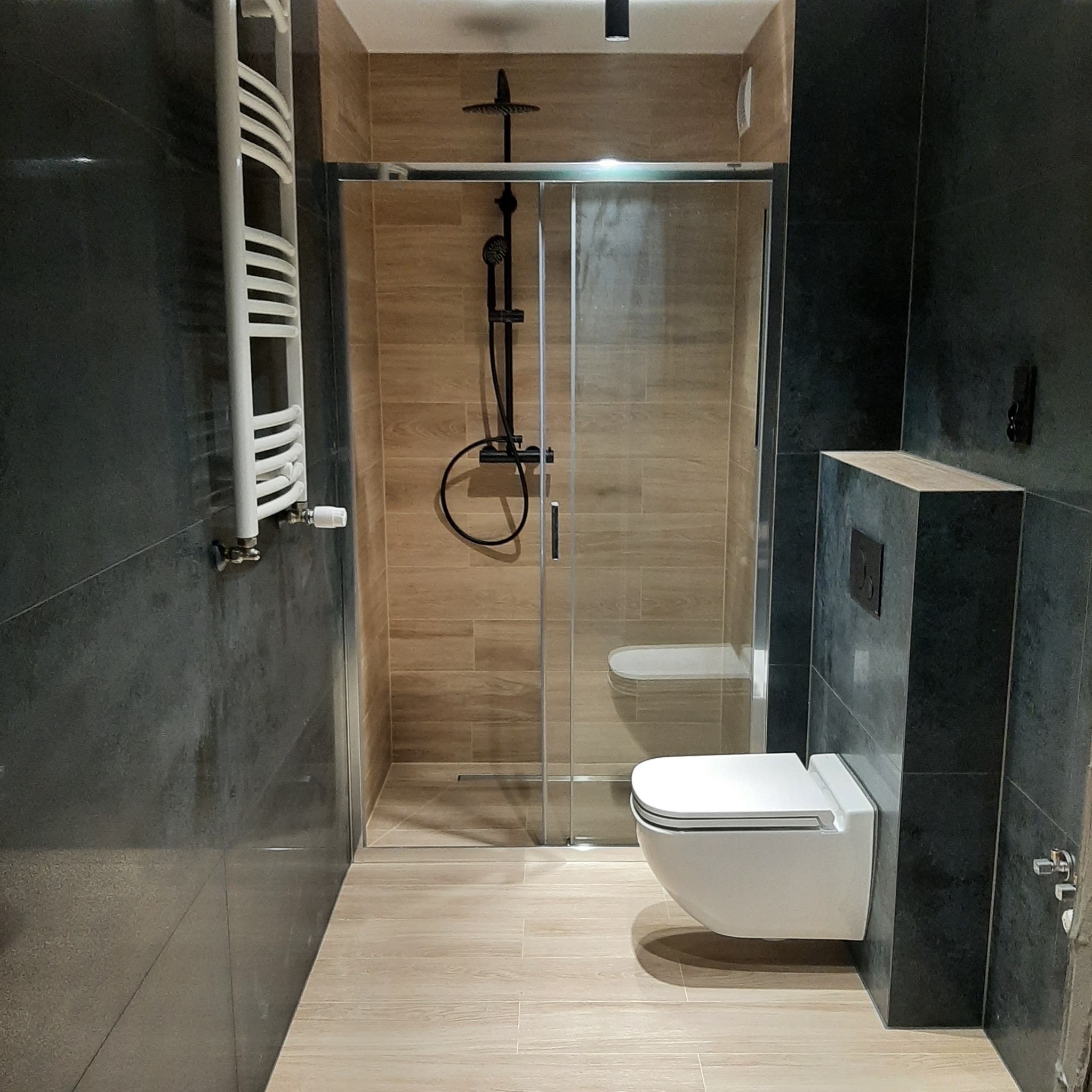 Nowoczesna łazienka z kabiną prysznicową typu walk-in, podwieszaną toaletą i grzejnikiem drabinkowym na ciemnoszarej ścianie, wykończona płytkami imitującymi drewno.