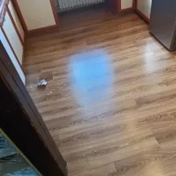 Jasna kuchnia po remoncie z nową podłogą z paneli laminowanych o wzorze drewna, widoczne zabezpieczone folią meble i okno, oraz fragment czarnej, marmurkowej podłogi w przejściu.