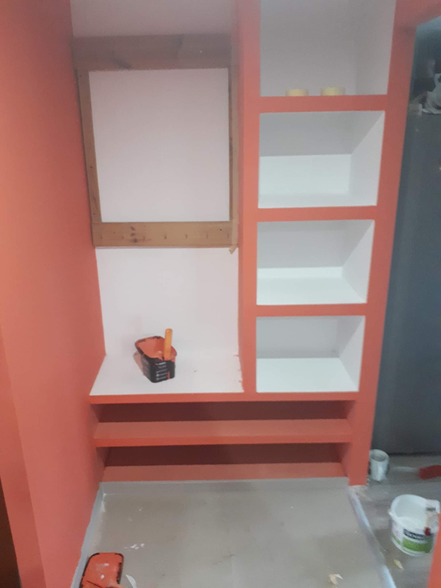 Wnęka z półkami w trakcie malowania na pomarańczowo, widoczne niedomalowane fragmenty i akcesoria malarskie.