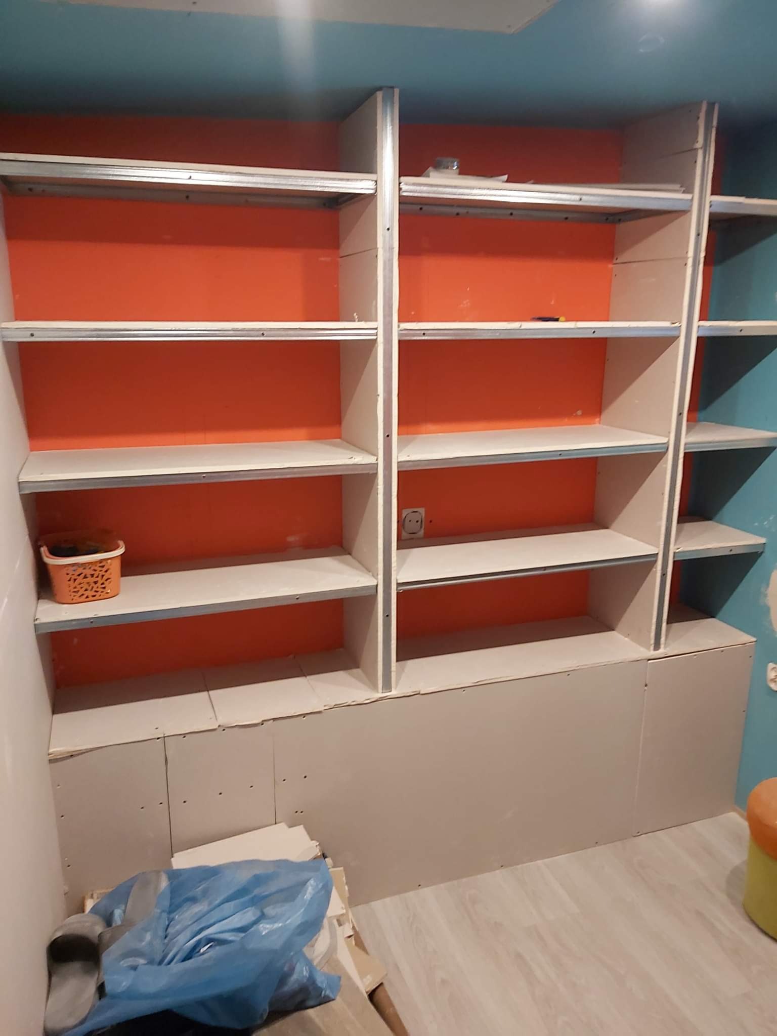 Konstrukcja regału z płyty gipsowo-kartonowej na metalowym stelażu, widoczne pomarańczowe wnętrze półek, niebieski sufit i ściana, na podłodze niebieski worek foliowy i resztki materiałów.