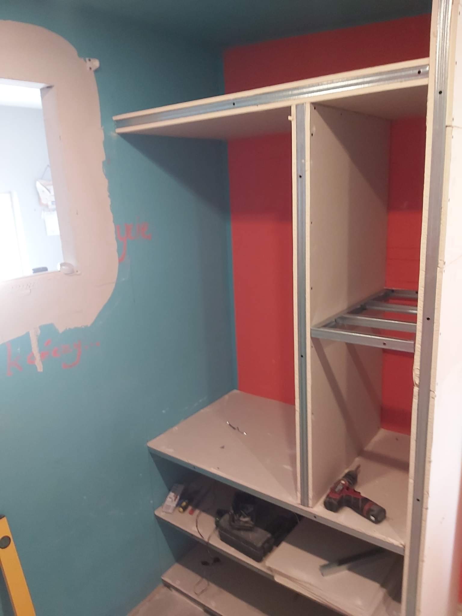 Wnętrze garderoby w trakcie budowy: szkielet z profili metalowych i płyt karton-gips, widoczne narzędzia na półkach, ściany w kolorze turkusowym i czerwonym w tle.