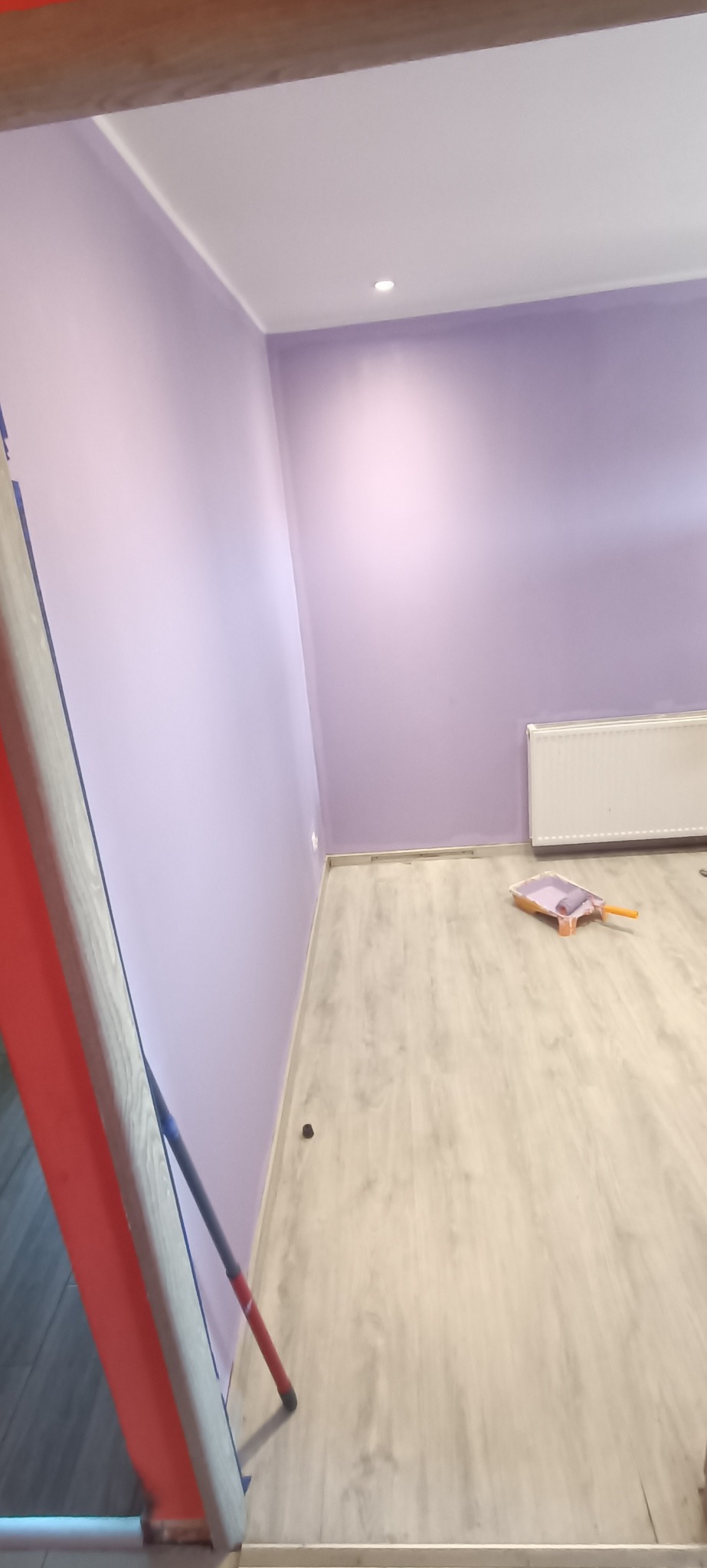 Świeżo pomalowane na fioletowo ściany pokoju z widocznym wałkiem do malowania na podłodze, grzejnikiem i fragmentem drabiny malarskiej.