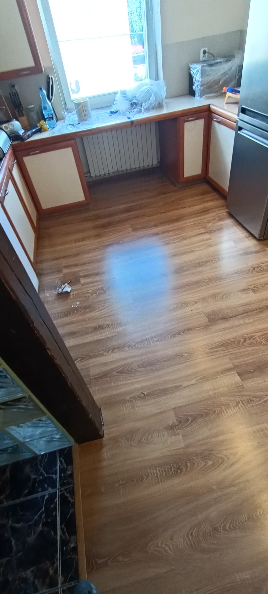 Jasna kuchnia po remoncie z nową podłogą z paneli laminowanych o wzorze drewna, widoczne zabezpieczone folią meble i okno, oraz fragment czarnej, marmurkowej podłogi w przejściu.