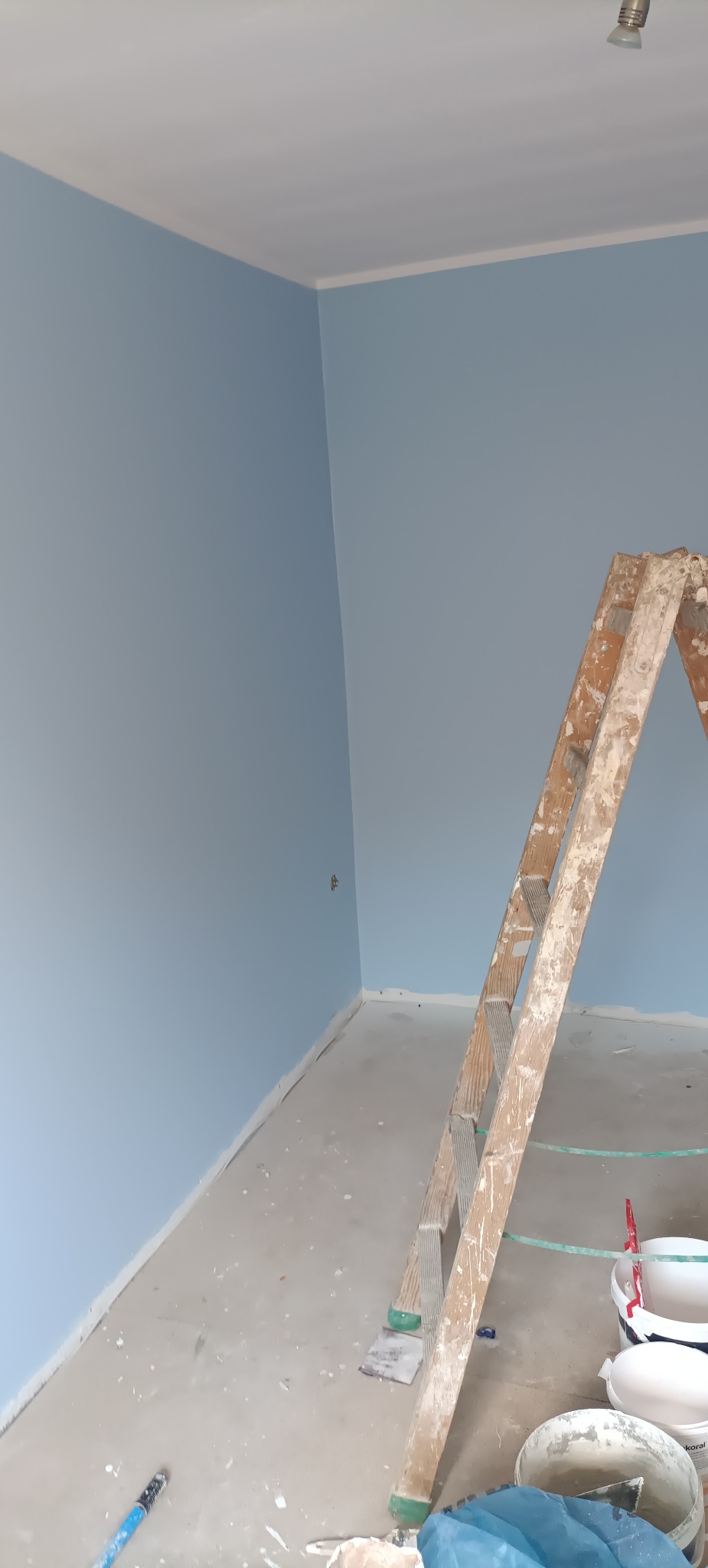 Pomieszczenie w trakcie malowania na jasny błękit, widoczna drewniana drabina, puszki z farbą i zabrudzona podłoga.