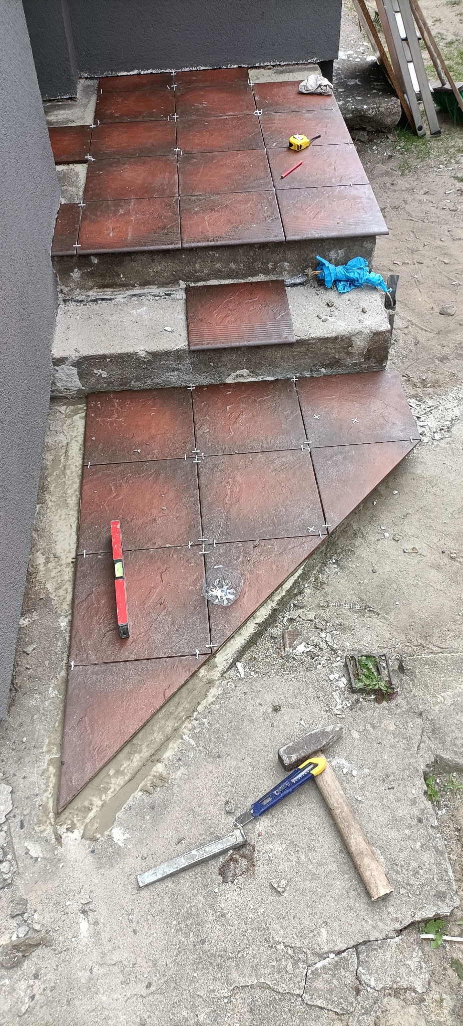Układanie płytek imitujących drewno na schodach zewnętrznych, widoczny poziomica, młotek, nóż i krzyżyki dystansowe.