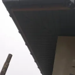 Fragment dachu z czarną blachodachówką, widoczna rynna i podbitka, ujęcie z dołu, w tle fragment ściany budynku i rusztowanie.