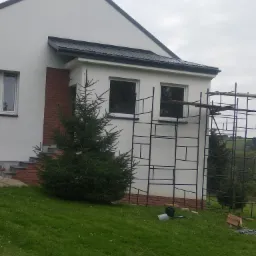 Biały dom murowany z elementami z cegły, z czarnym dachem, w trakcie budowy, widoczne rusztowania, na pierwszym planie mała choinka