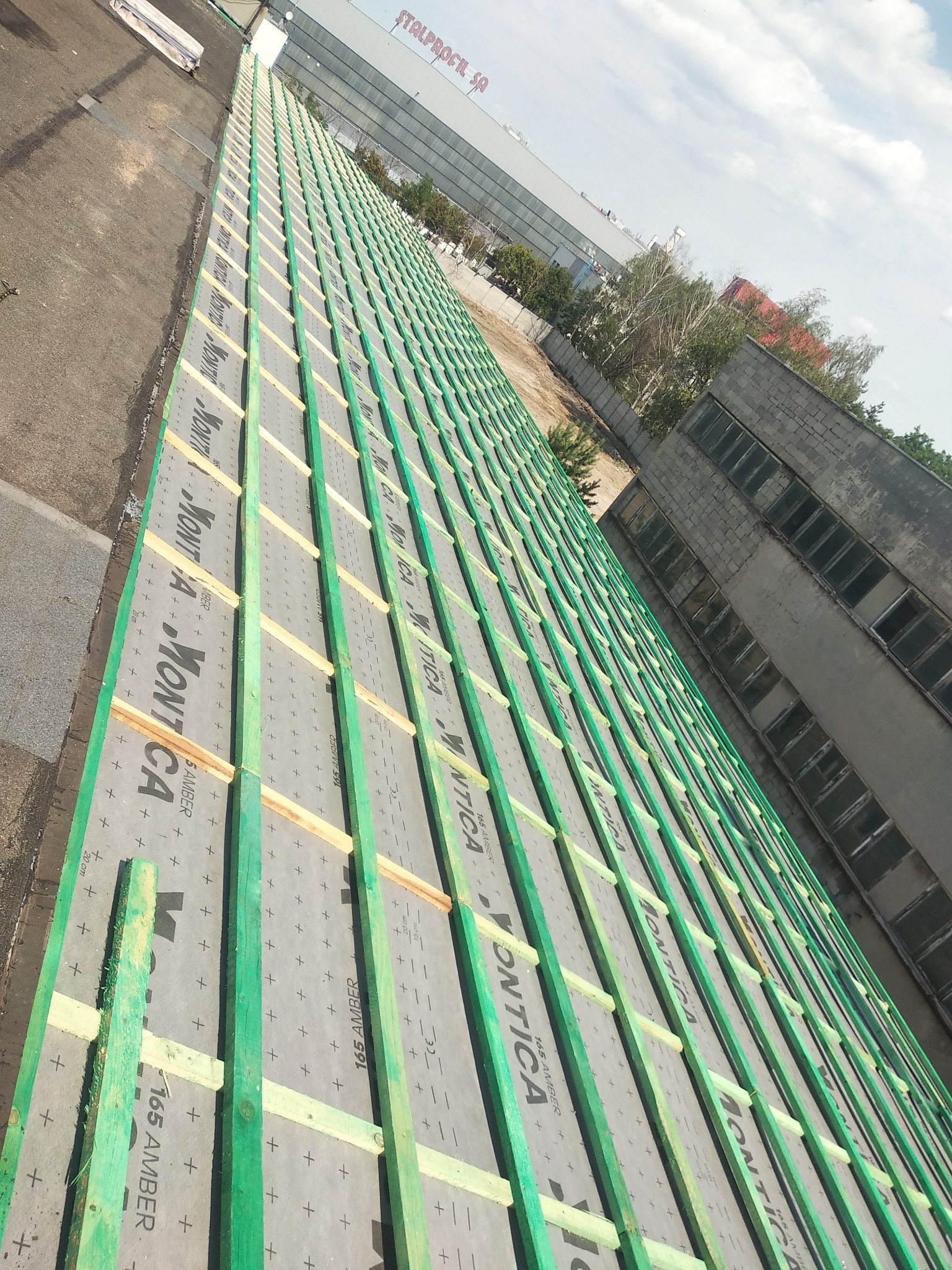 Konstrukcja dachu z łatami i membraną Montica 165 Amber, widok z góry w trakcie budowy, z budynkiem przemysłowym w tle.