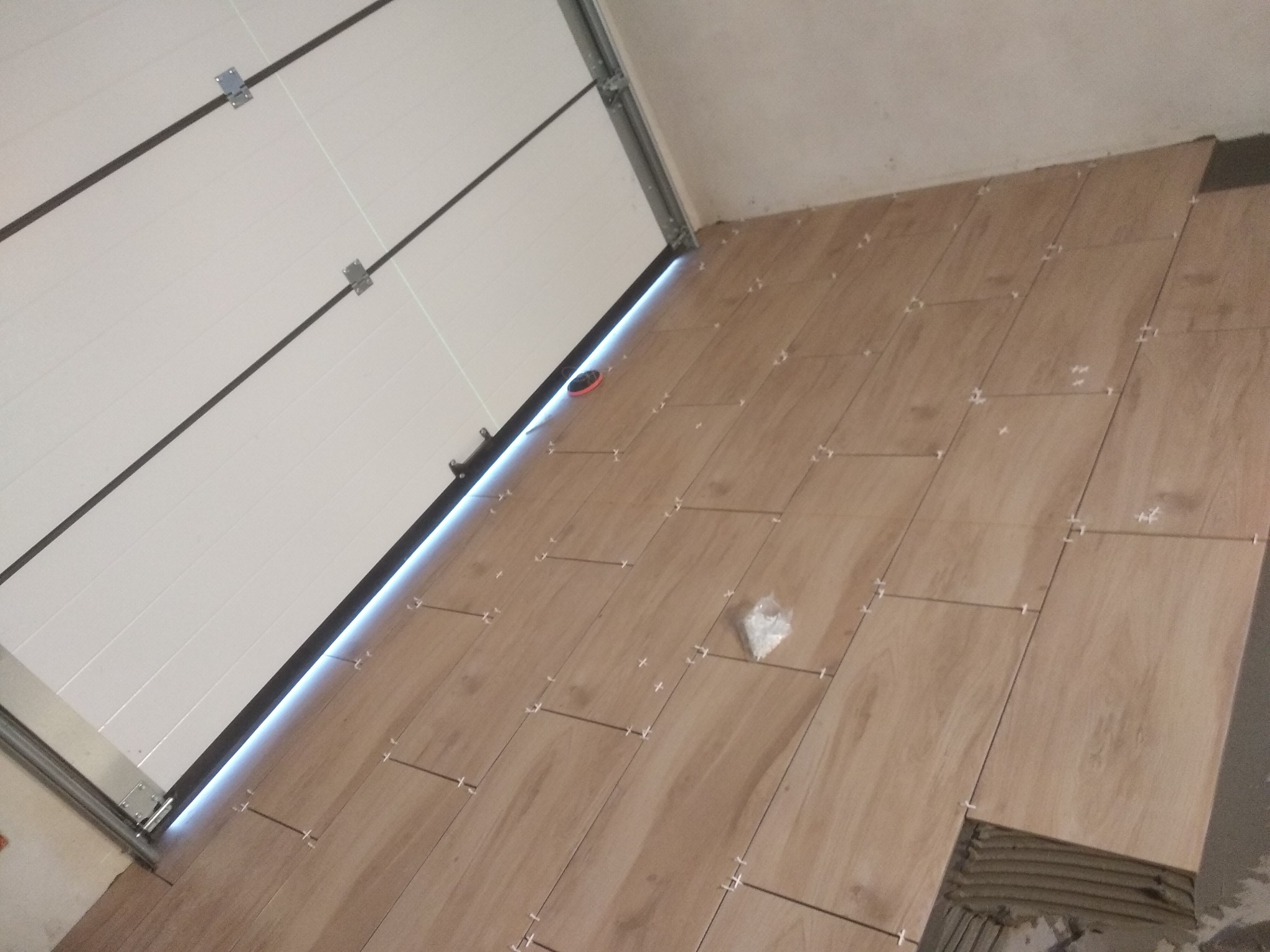 Układanie podłogi z płytek imitujących drewno w garażu, widoczna brama garażowa i krzyżyki dystansowe między płytkami.