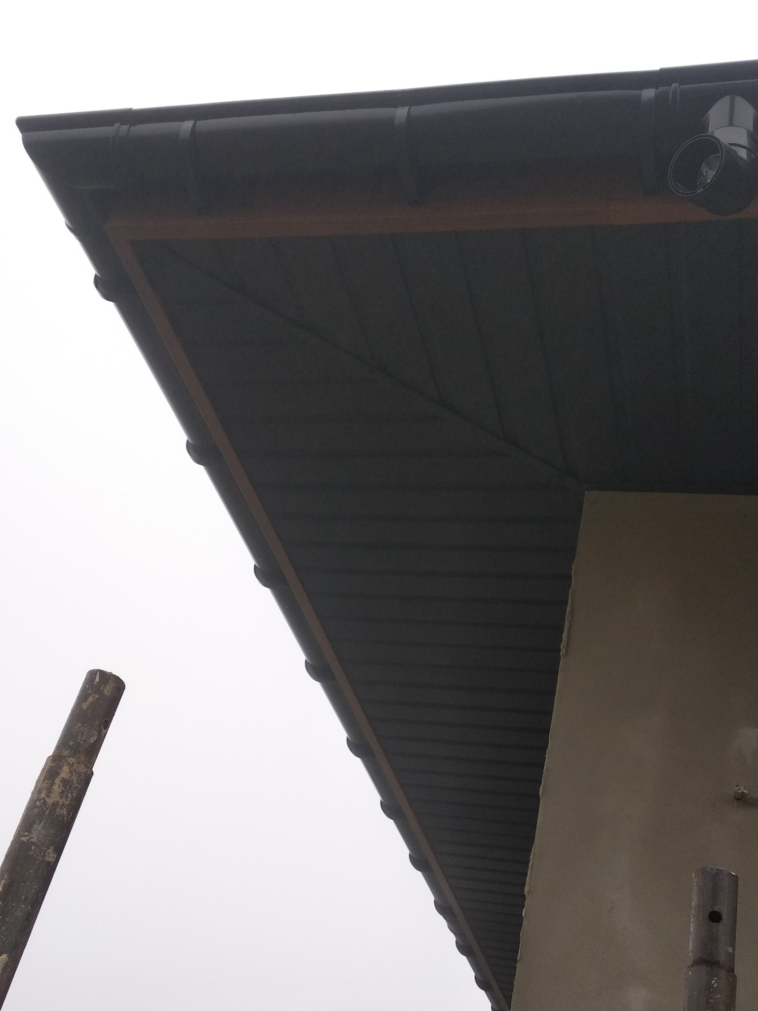 Fragment dachu z czarną blachodachówką, widoczna rynna i podbitka, ujęcie z dołu, w tle fragment ściany budynku i rusztowanie.