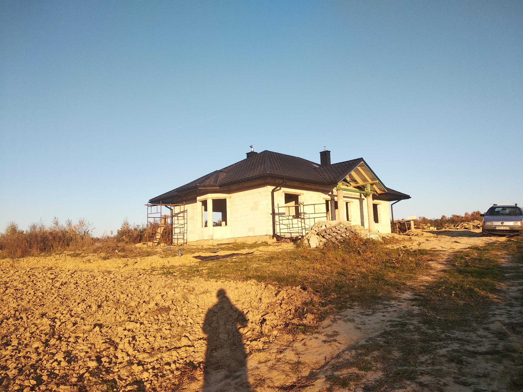 Surowy stan domu jednorodzinnego z szarej cegły, pokrytego ciemnym dachem, z widocznymi rusztowaniami i stertą materiałów budowlanych obok, na tle nieba i zarysu krajobrazu.