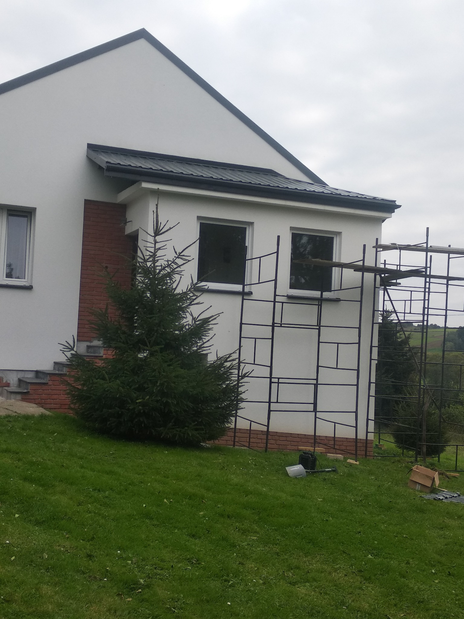 Biały dom murowany z elementami z cegły, z czarnym dachem, w trakcie budowy, widoczne rusztowania, na pierwszym planie mała choinka