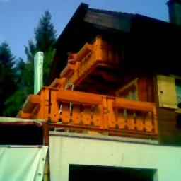 Drewniany balkon w trakcie montażu, widoczne elementy konstrukcyjne i zabezpieczona rura, tło z drzewami iglastymi.