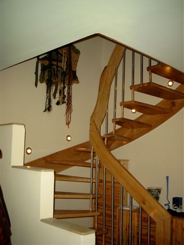 Spiralne schody z jasnego drewna z metalowymi balustradami, oświetlone punktowymi lampami w ścianie, z dekoracją wiszącą na ścianie.