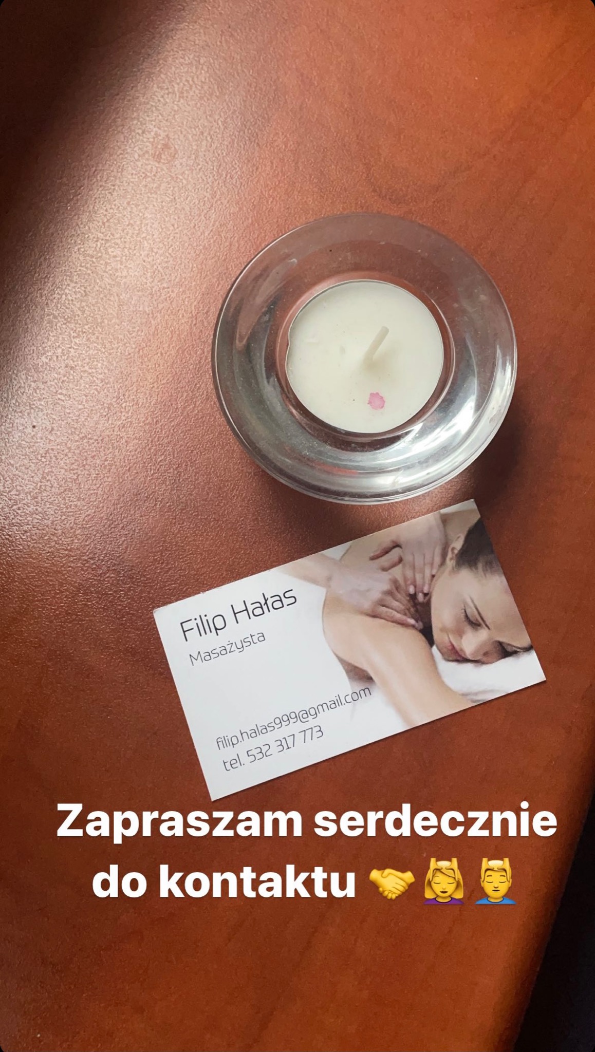 Na brązowym tle leży wizytówka z imieniem Filip Hałas, tytułem Masażysta i zdjęciem kobiety podczas sesji. Obok wizytówki stoi świeczka w szklanym świeczniku.